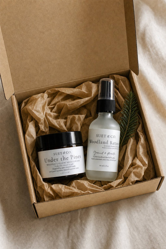 A Suet & Co gift set in a brown kraft box, featuring a jar of 'Under the Pines' whipped tallow moisturizer and a bottle of 'Woodland Retreat' shower & room spray, arranged on brown paper filler with a small pine sprig, on a neutral fabric background.