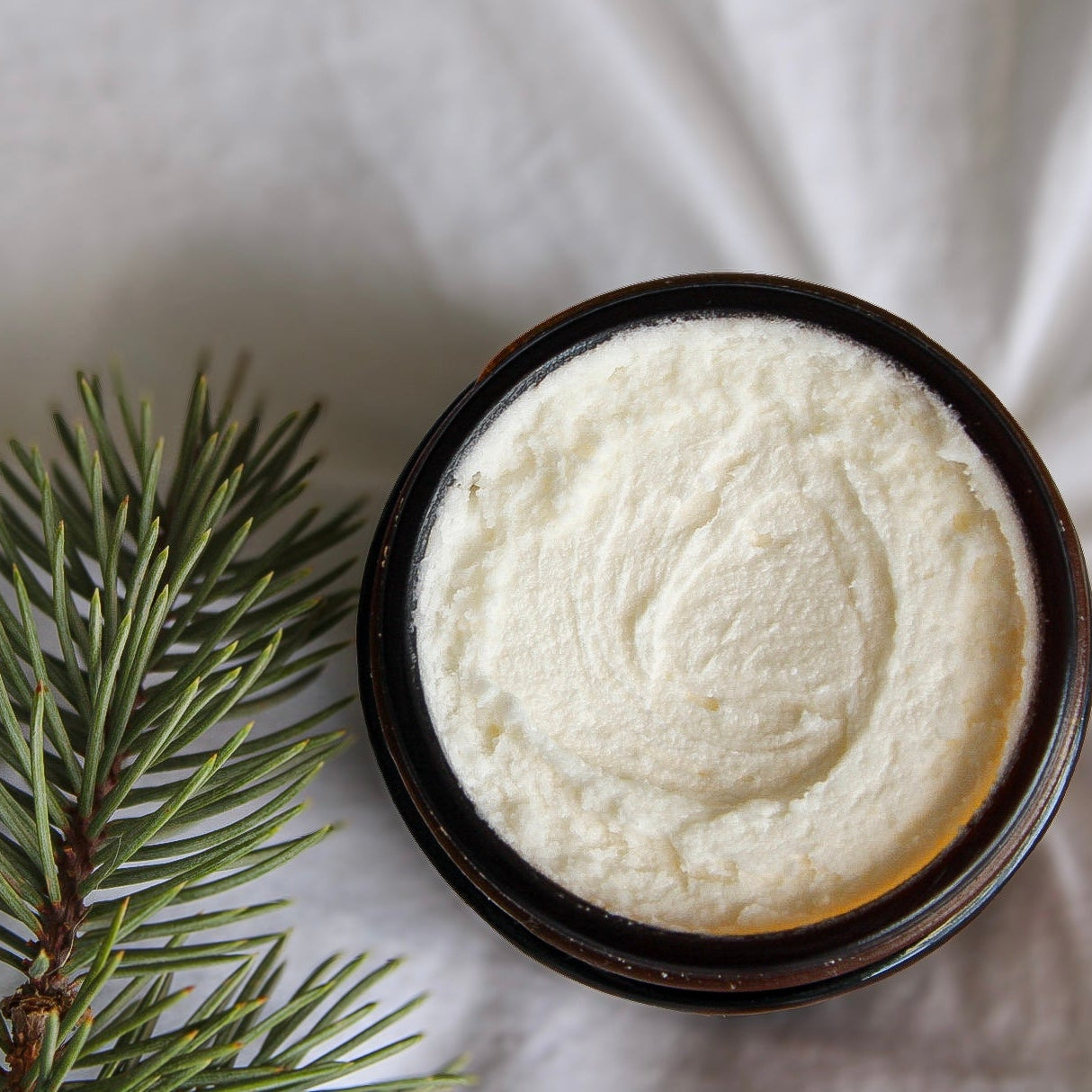 Container of Under the Pines whipped tallow moisturizer with a sprig of pine on white fabric background