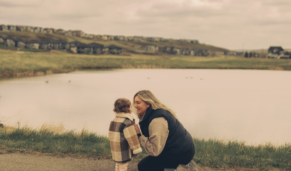 A woman kneels beside a child near a calm lake, with grassy hills and houses in the background under an overcast sky.