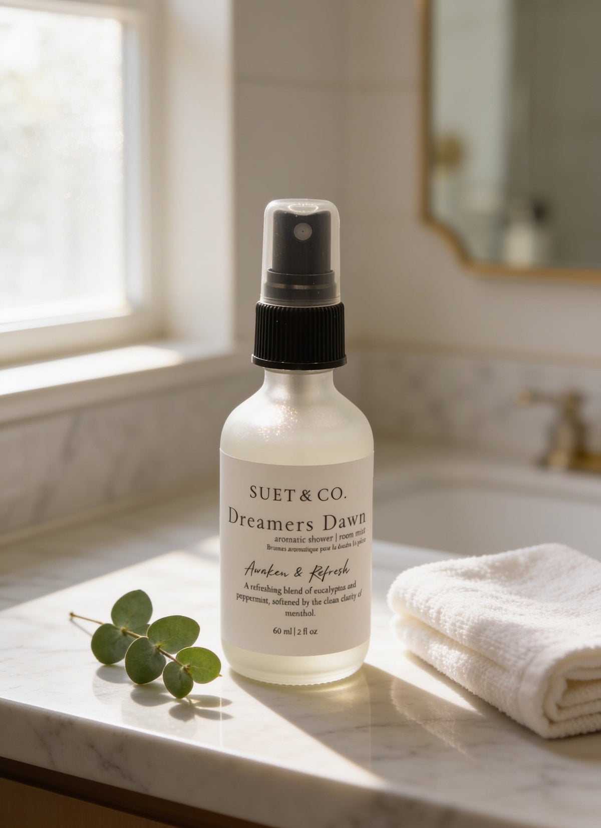 A frosted glass spray bottle labeled 'SUET & CO. Dreamers Dawn' stands on a marble bathroom counter beside a folded white towel and a small green sprig, with soft sunlight and a window in the background.