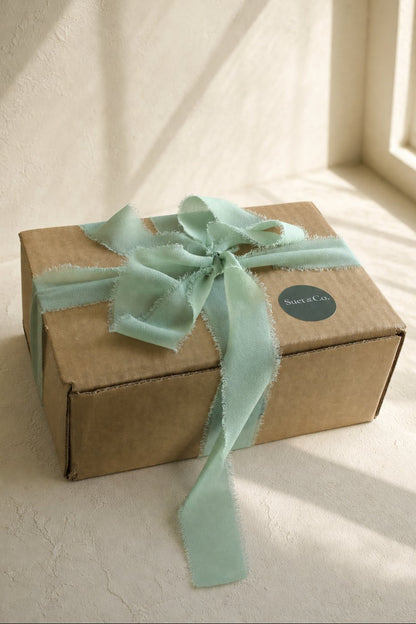 A brown cardboard gift box tied with a pale mint green satin ribbon and bow, placed on a light textured surface near a window with soft natural light.