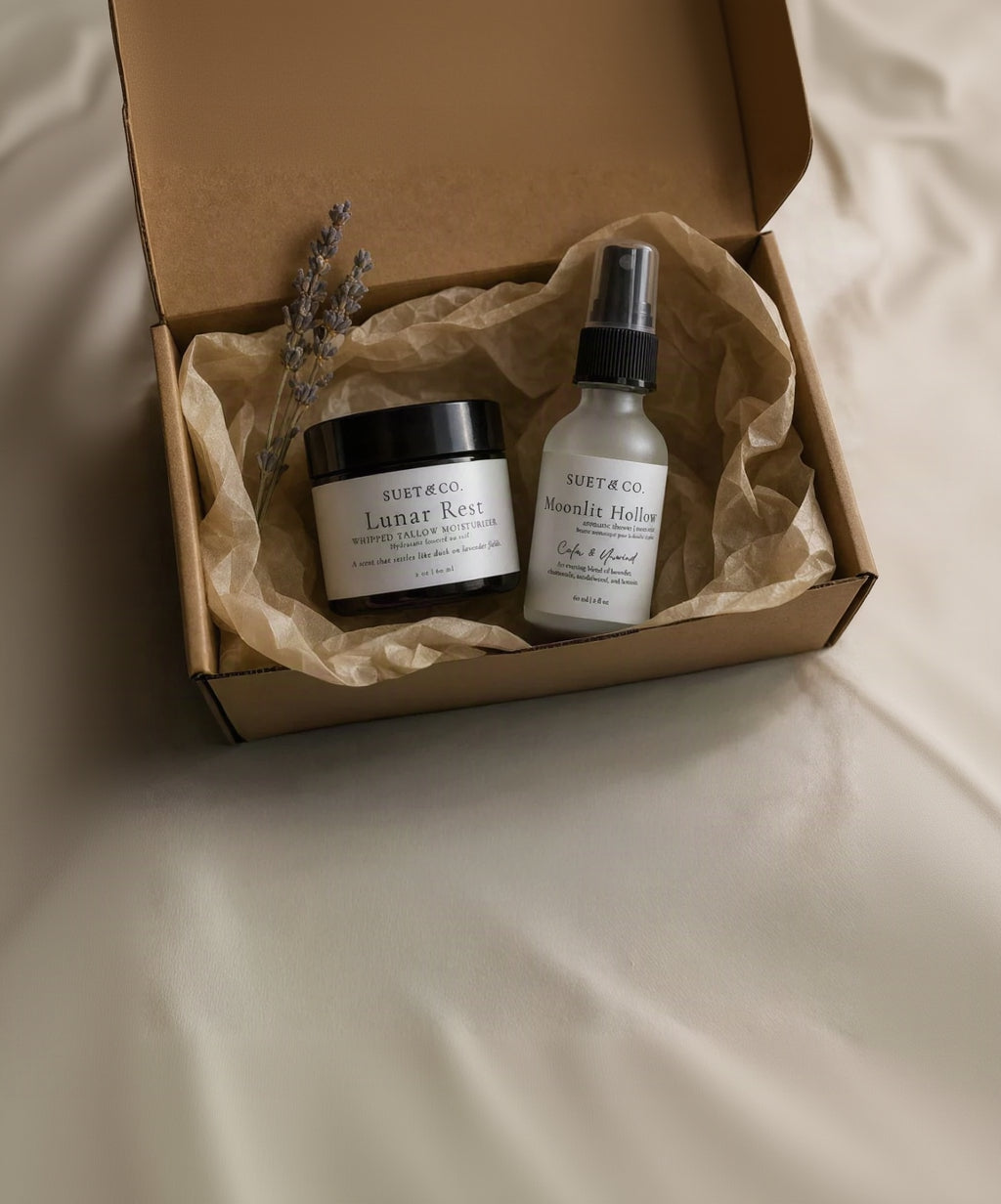 A Suet & Co. gift set in a brown cardboard box lined with brown tissue paper, featuring a black jar of 'Lunar Rest' body tallow and a frosted white spray bottle of 'Moonlit Hollow' aromatic mist, styled with dried lavender sprigs on a light beige fabric background.